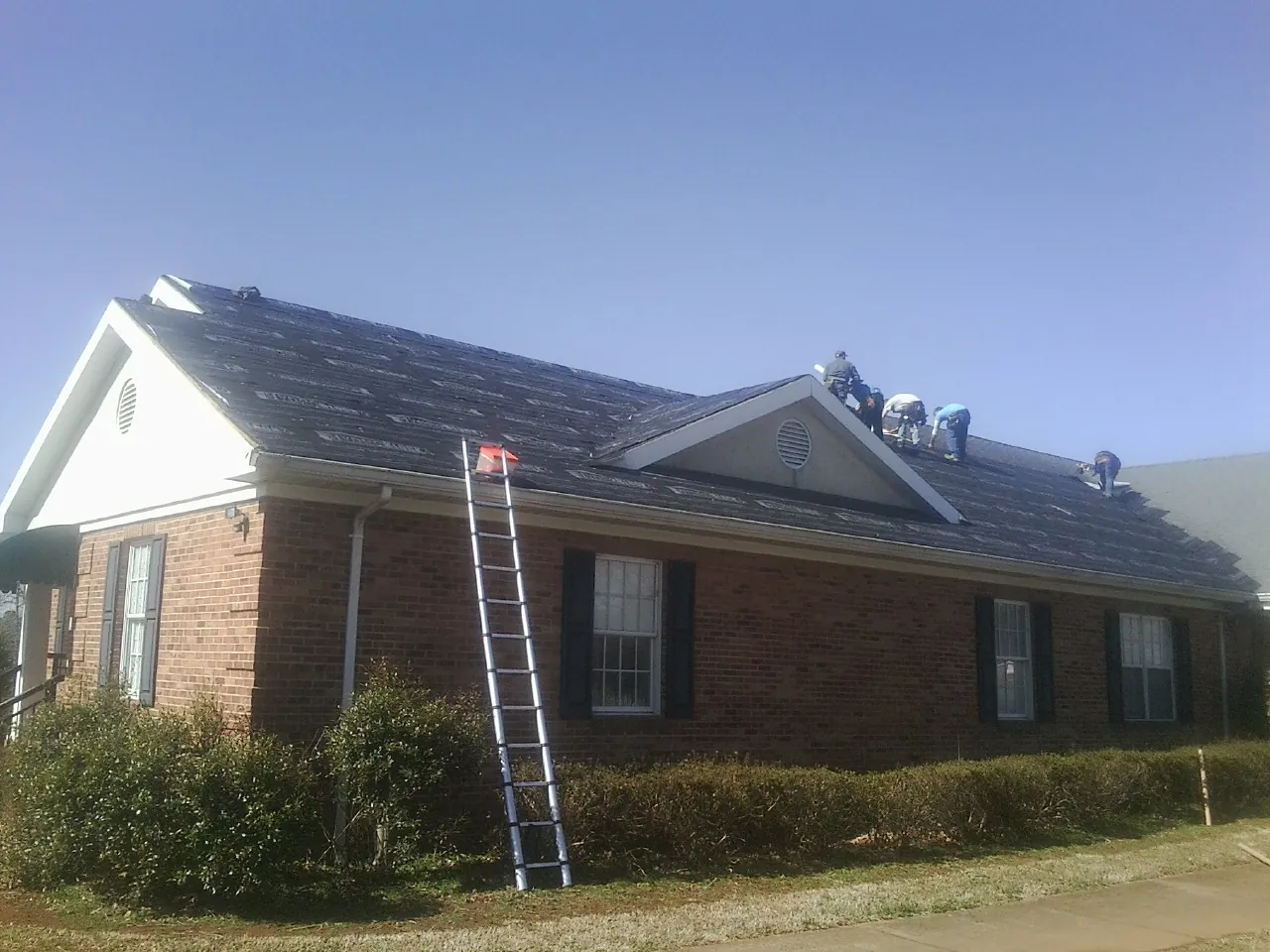 Crew tearing shingles off a brick ranch roof with a ladder set — Metal Roof Repair work by our crew in Spiritwood