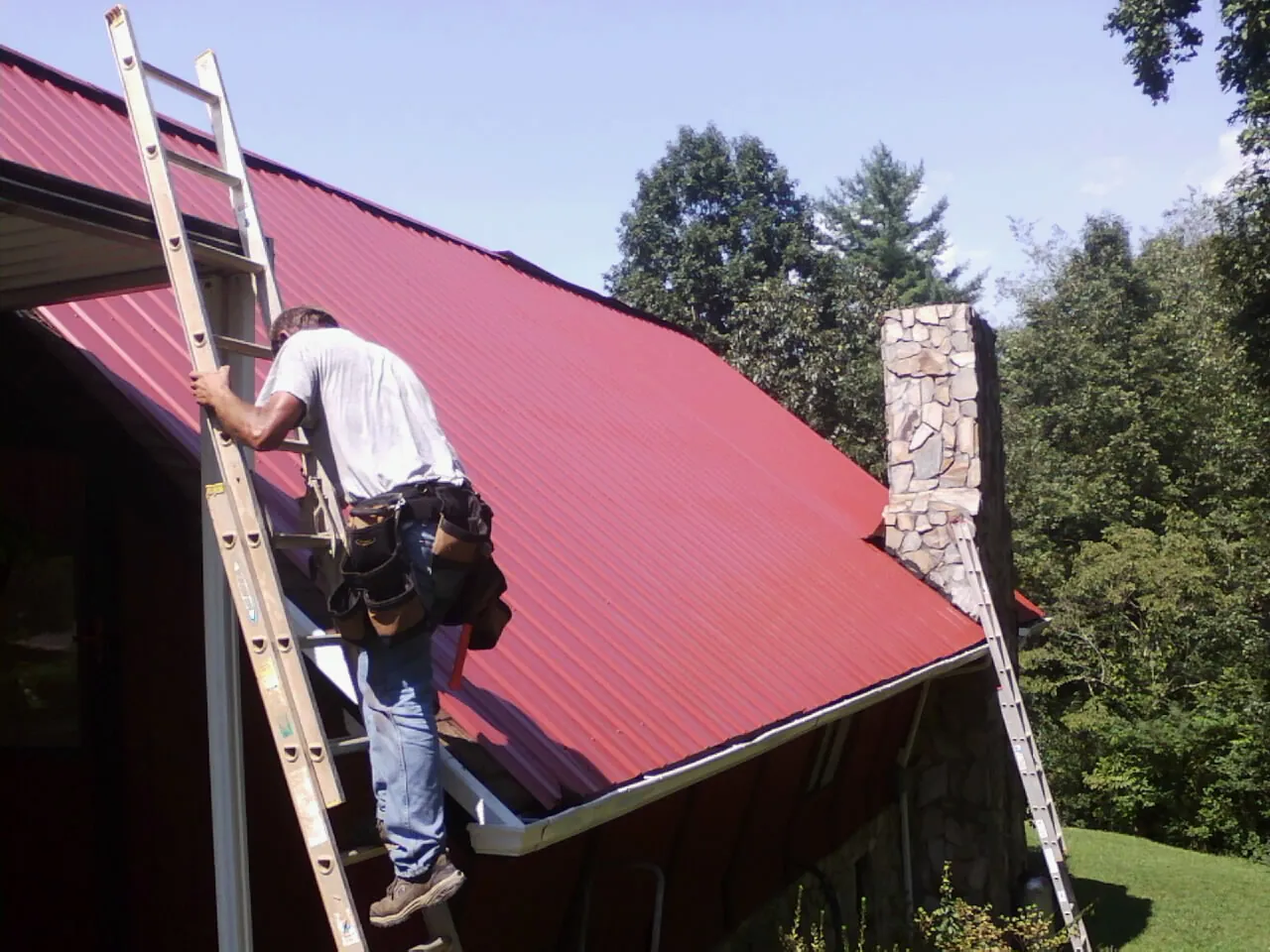 Roofer on a ladder beside a red metal roof and stone chimney — Soffit & Fascia Repair by Roofing Brothers in Spiritwood